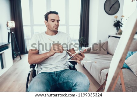 Portrait of Disabled Young Man Painting Picture. Handsome Person Sitting in Wheelchair Opposite Easel Holding Paintbrushes in Hands Wearing Casual Clothes While Sitting in Living Room at Home