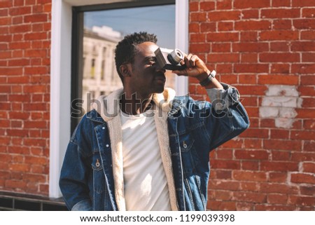 Fashion african man photographer with vintage film camera taking picture on city street, wearing jeans jacket, brick wall background