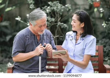 Nurse with patient sitting on bench together looking at tablet. Asian old man and young woman sitting together talking. Relax mood.