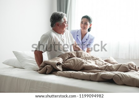 Senior man in bed trying to get up and nurse helping him. Old asian man and beautiful asian nurse woman in bedroom and open curtain. Senior home service concept. Royalty-Free Stock Photo #1199094982