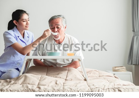 Young attractive nurse feeding breakfast to senior man in bed. Old asian male with white beard with young attractive asian woman caretaker. Senior home service concept. Royalty-Free Stock Photo #1199095003