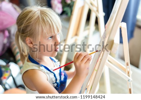 Cute blond smiling girl painting on easel in workshop lesson at art studio. Kid holding brush in hand and having fun drawing with paints. Child development concept