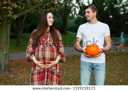 Happy young and stylish pregnant couple wife in checked dress and husband in white t-shirt posing, having fun in park with orange pumpkin. Positivity and romantic family preparing for Halloween.