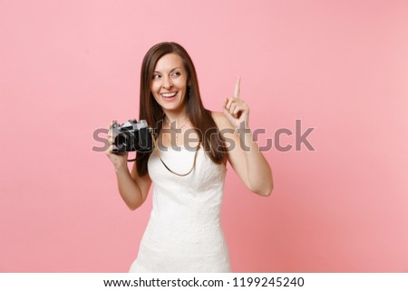 Happy bride woman in wedding dress pointing index finger up holding retro vintage photo camera choosing staff photographer isolated on pink background. Wedding to do list. Organization of celebration