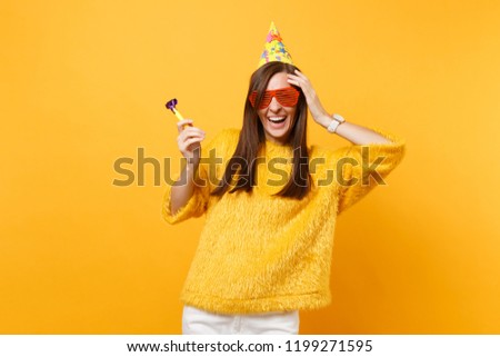 Laughing woman in orange funny glasses, birthday hat with playing pipe putting hand on head celebrating isolated on yellow background. People sincere emotions, lifestyle concept. Advertising area