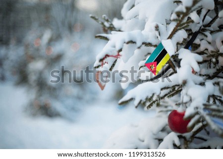 Mozambique flag. Christmas background outdoor. Christmas tree covered with snow and decorations and Mozambique flag. New Year / Christmas holiday greeting card.