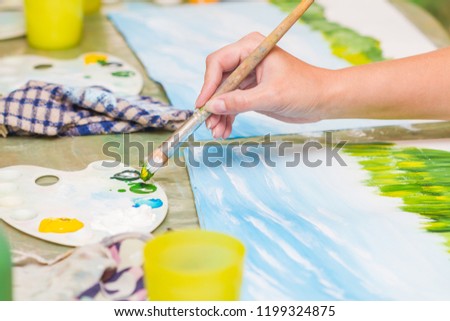 A girl paints a picture by gouache. A dirl drawing at white paper. The hand and paint brush. Closeup, selective focus