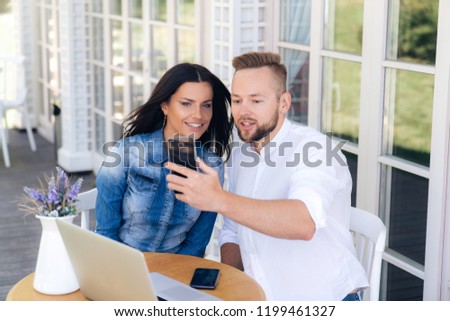 Close-up of a beautiful girl with dark hair and a stylish guy sitting in a cafe outdoors, chatting on a video call with a friend, smiling and making an appointment.