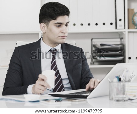 Male is working at a computer and drinking coffee in the office.