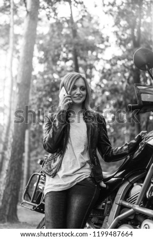 Portrait of happy woman talking on phone,, black and white, BW, dressed in a leather jacket and jeans, with adventure tourist motorcycle, in the forest, autumn day, off road travel vertical photo