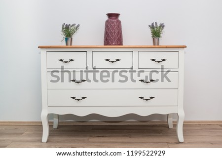 White wooden dresser with three vases and flowers on white wall background. Chest of drawers close up. Royalty-Free Stock Photo #1199522929