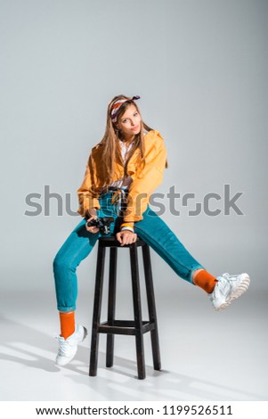 smiling stylish photographer holding camera while sitting on stool on grey