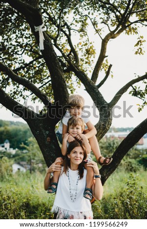 Charming young mother hugs tender her little sons standing under old tree in the rays of evening sun