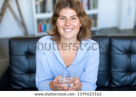 Front view portrait of a happy teen holding a glass of water looking at you sitting on a couch in the living room at home