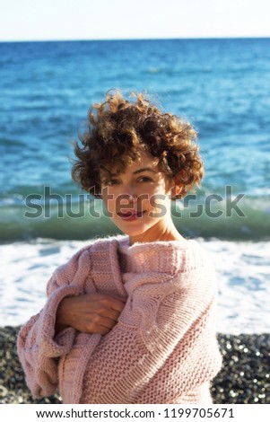 A lovely, short-haired, curly-haired, brunette, young woman in a rose sweater in the backdrop of the sea Royalty-Free Stock Photo #1199705671
