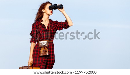 Beautiful girl in plaid dress with bag and binocular on sky background