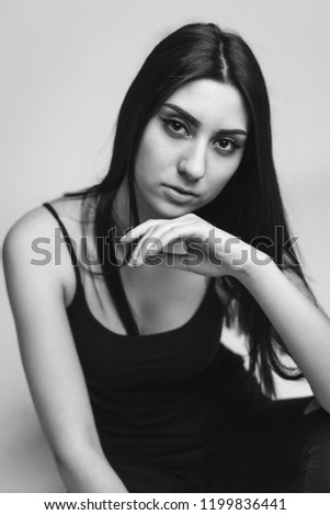 Close up portrait of beautiful caucasian woman with long brunette hair in black clothes posing at the camera on grey background. Black and white photo