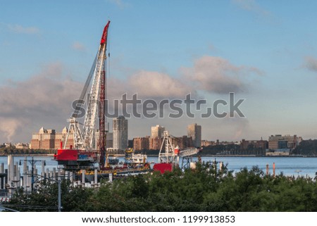Two red painted cranes are sitting on the water of the Hudson River. A building stands in front of the cranes. The New Jersey shore can be seen in the distance. Pink clouds and blue is above.