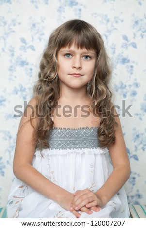 Portrait of sincere cheerful tender young blond girl child with grey eyes and wavy long hair in sitting posture looking at camera