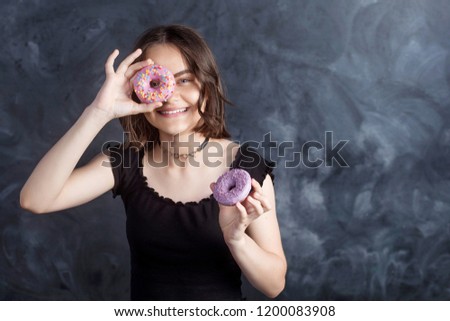 Portrait of joyful girl covering her eyes with donuts on black background. Attractive girl holding fresh donuts and having fun with sweet-stuff. Good mood, diet concept.