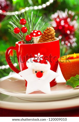 Picture of luxury Christmastime table decoration, red coffee cup decorated with Santa Claus star, yellow candle and berry twig, beautiful white dinnerware on Christmas tree background, New Year party