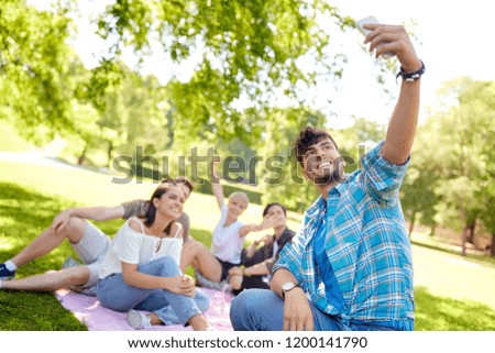 friendship, leisure, technology and people concept - group of friends chilling on picnic blanket abd taking selfie by smartphone at summer park