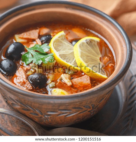 Traditional Russian soup Solyanka with meat, sausages, vegetables, capers, pickles and olives with lemon, seasonings and spices. Served with sour cream. Rustic style.  Selective focus, Square picture