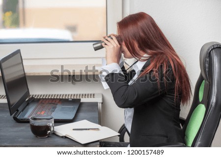 young professional woman sitting in the office in front of a laptop, looking at through binoculars, she is focused, looking for something