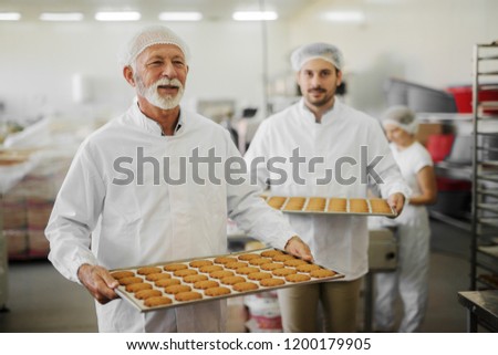 Picture of two male food factory employees in sterile clothes holding trays full with fresh cookies. Working on production line in food factory.Smiling and working together.