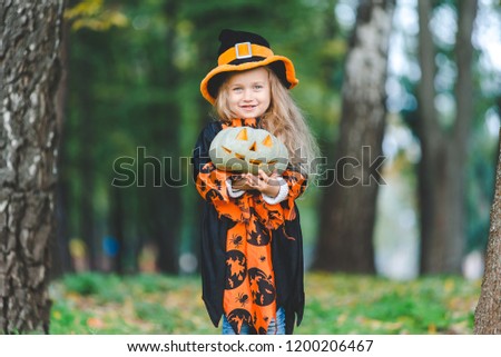 Cute little girl in witch costume for the holiday of Halloween. She holds a pumpkin in her hands and stands in the park.