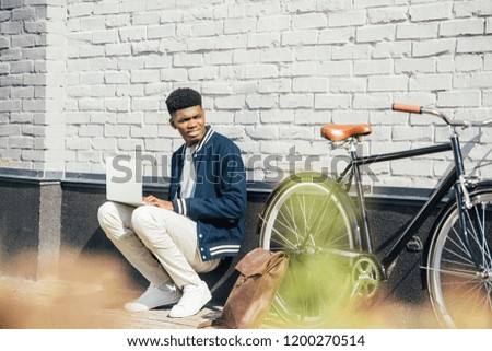 handsome african american freelancer working with laptop near bicycle at white brick wall