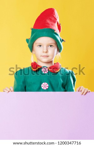 Little girl in a Christmas elf costume on a yellow background. Beautiful child in a red hat with bells. Copy space on a pink background.