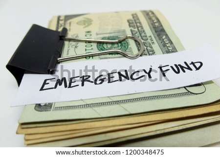 Closeup of US dollars in paper clip on white background with note written EMERGENCY FUND : Concept of setting money saving goal for rainy day.  Royalty-Free Stock Photo #1200348475