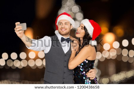 celebration, people and holidays concept - happy couple in santa hats taking selfie by smartphone over christmas tree lights background