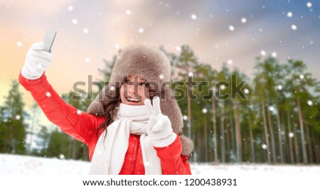 people, technology and leisure concept - happy smiling woman in winter fur hat taking picture by smartphone on selfie stick over winter forest background and snow