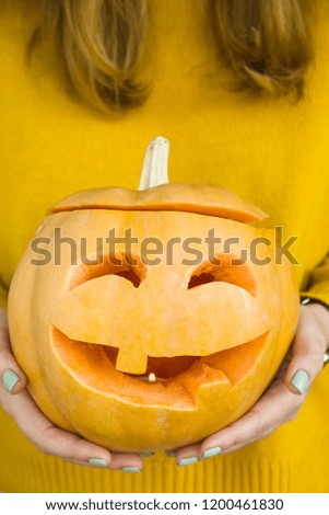 A festive pumpkin with eyes carved and a mouth with a candle inside in the hands of a woman in a yellow sweater. Halloween decoration concept