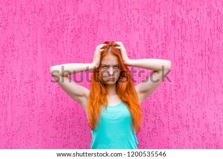 Portrait of frustrated redhead girl holding hands on head and looking at camera over pink background