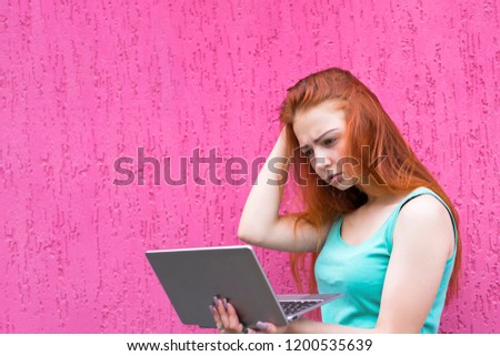 Young confused lady standing over pink wall background using laptop computer. Red hair girl using tablet pc. Copy space in left side