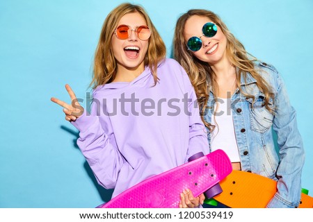 Two young stylish smiling blond women with penny skateboards. Models in summer hipster sport clothes posing near blue wall in studio. Positive girls going crazy Royalty-Free Stock Photo #1200539419