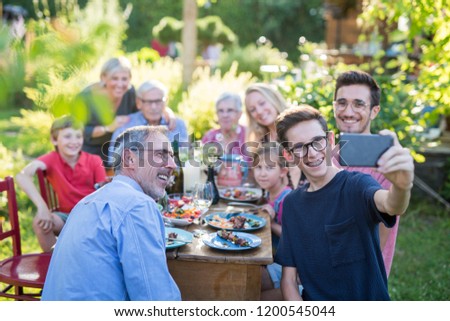 In the summer, a family of three generations having fun around a table in the garden sharing a meal. A teenager does a selfie with all the guests