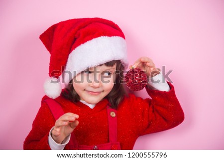 Cute smiling little girl in Santa hat and costume holding red Christmas ball in studio on pink background. New Year banner with empty space.