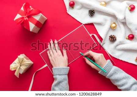 Female hands, girl wrote in a Christmas card, red table with gift boxes for Christmas and New year, green pensil, background with copy space, for advertisement, top view