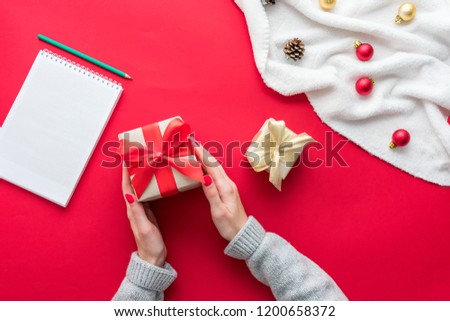 Female hands, girl holds a gift, white Notepad, red table with gift boxes for Christmas and New year, green pensil, background with copy space, for advertisement, top view