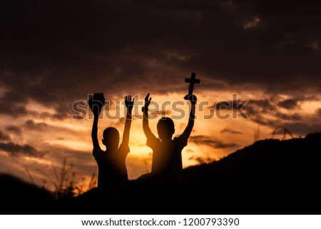 Two children worship God by holding christian Cross and Bible with light sunset background,christian concept.