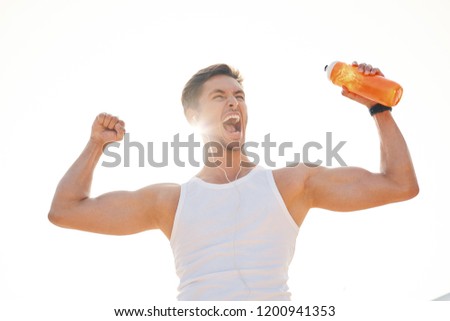 Athletic muscular man feeling victory and enjoys success holding a bottle of water. Emotion, power, gesture - young man celebrating victory and screaming with sun light in background.