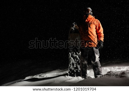 Snowboarder in orange sportswear and mask standing with a board on a snowy hill at night
