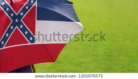 Mississippi state flag umbrella. Closeup of a printed umbrella over green field/lawn background. Rainy weather / climate change and global warming concept.