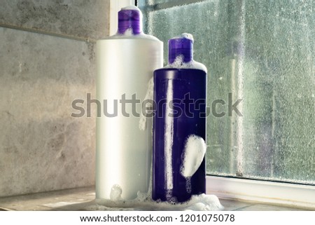 White and purple shampoo and conditioner bottles with bubbles running down side on interior bathroom window sill, grey tiled background to left of image, frosted class with bottle reflection to right.