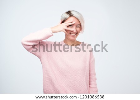 Young hipster woman in pink sweater peeping at the camera through her fingers. Picture of playful female looking through her hands covering her face on gray background