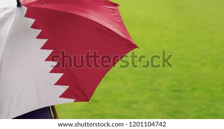 Qatar flag umbrella. Closeup of printed umbrella over green grass field background. Landscape, side view. Rainy weather/ climate change and global warming concept.
